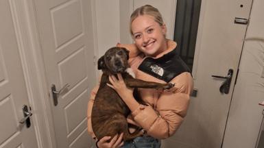 A young woman is holding a dog while smiling at the camera in a house, infront of the front door.