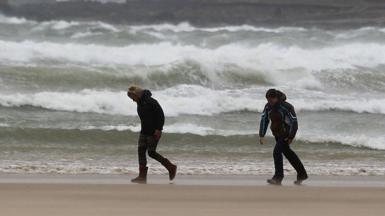 Two people bend into the wind as they walk along a beach with crashing waves behind them