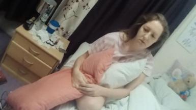 A woman with brown hair and wearing a pink top clutches a pillow while sitting on a bed. Behind her a teddy bear can be seen on a chair.