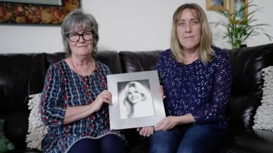 Wendy Ramsay and daughter Emma, sitting on a leather sofa in a living room holding a picture of Samantha. Wendy has grey hair and glasses and wears a blue and red top, while Emma is blonde and wearing jeans with a navy top with a pattern of small stars.