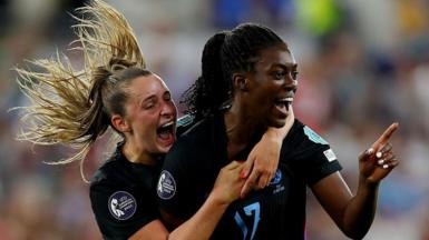 Two England players with faces full of joy and wide smiles, both wearing dark blue short sleeve team shirts. With one's blonde pony tile flying wildly. 