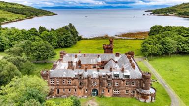 An aerial view of the red sandstone lodge with its ornate turrets and battlement-like rooftops. The lodge is set in an grounds with large areas of grass and trees. In the background is the sea, with a few boats dotted around a bay, and mountains in the distance.