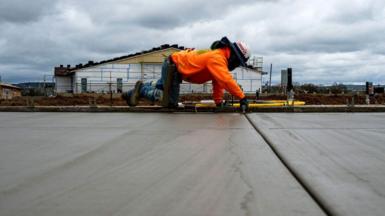 A contractor in a bright orange sweatshirt, jeans and construction hat is on hands and knees smoothing concrete flooring at the Toll Brothers Redwood, 