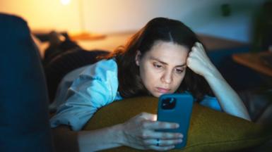A young women is lying on a bed staring at her phone in a dark room, there is a table lamp in the background and she is leaning on a cushion