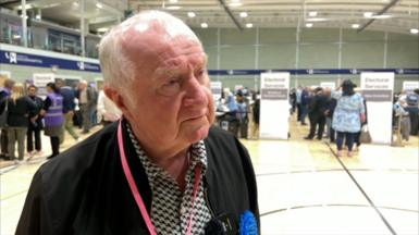 A grey haired man in an election counting venue. he's wearing a black jacket with black and white patterned shirt