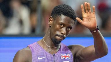 Former Great Britain sprinter Reece Prescod waves with his left hand as he wears purple and white kit with a union jack badge and Nike swoosh logo on the front