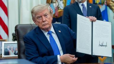 Trump, wearing a blue suit and sitting at desk in the Oval Office, holding up a signed executive order