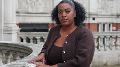 Nadine is standing outside, looking at the camera. She has black curly hair and is wearing a brown cardigan.
