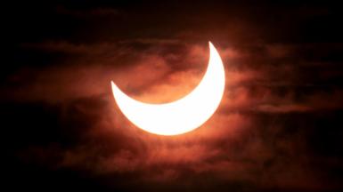 A partial solar eclipse showing the bright crescent of the sun as the moon moves across it from the top left corner. The sky is dark with clouds visible behind the sun