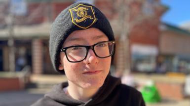 Johnpaul looks directly at the camera with a blurred-out building in the background. He is wearing black glasses, a black hoodie and a black beannie hat with a crest on it.