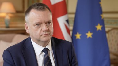 Nick Thomas-Symmonds sits for an interview, wearing a dark suit, white shirt and tie. UK and EU flags are positioned behind him.