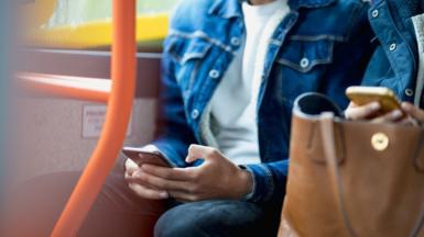 A man in a white t-shirt and blue denim jacket sits on a bus next to the window and uses his phone. His face is out of the camera shot. Another passenger sat next to him also uses their phone.
