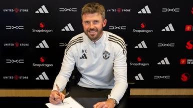 Michael Carrick sat down in Manchester United training kit, posing holding a pen over a contract document