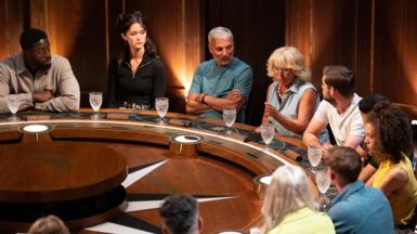 Traitors from the BBC TV series 4 sit at the round table. The focus is on Traitor Fiona who is speaking to the rest of the group