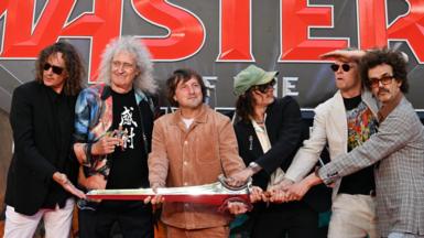 Five men standing next to each other holding a silver sword prop, in front of a movie premier sign with bold red writing 