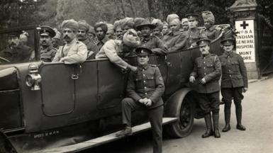 In a black and white photo from 1914 or 1915, a large motor vehicle carries around two dozen Indian soldiers. Three British officers stand beside the vehicle. In the background, a sign reads: "No. 8A Indian General Hospital, Mont Dore".