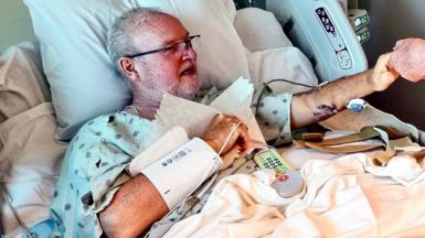Peter Smith lies in a hospital bed holding out his hand to touch another person's (not pictured) hand, he's wearing glasses and hospital gown with medical equipment around him.