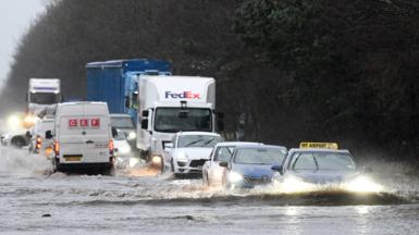 Cars drive through heavy floods on the roads in Northern Ireland. There are several cars and lorries driving through deep water. They hae their headlights on as the light is low