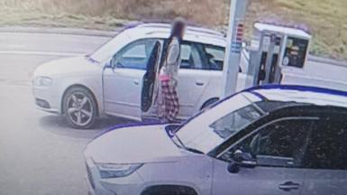CCTV footage showing a thief wearing red and white pyjama trousers and a grey jumper filling up the tank of their silver car. Their face is blurred out. There's another car parked up next to them in the forecourt.