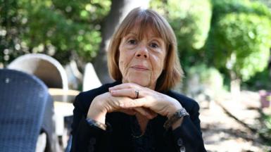 A woman with short brown hair wearing a dark blue jumper sits in a sunlit garden. Gisèle Pelicot's hands are crossed under her chin and she looks into the camera