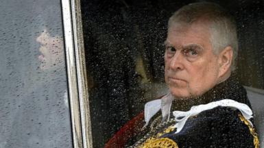 Andrew sitting in a car looking out the window which is covered in rain. He wears a fancy, royal looking outfit and looks glum. A woman sits next to him in the car. 