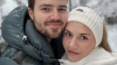 Two people standing close together in the foreground, wearing padded winter jackets, with one in a dark green coat and the other in a white coat and knitted headband. Snow is visible on the clothing. Behind them are snow-covered trees and branches.
