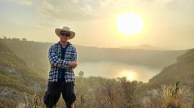 Gus in Mexico, standing on a hill with his arms crossed in a chequered overshirt, sunglasses and a wide-brimmed sun hat with a lake and low sun in the background