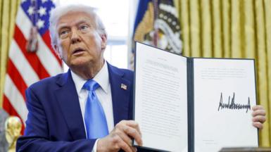 Donald Trump in Oval Office holds up folder displaying a signed executive order