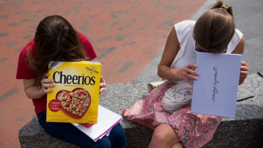 Two girls look into cardboard boxes, one a cereal box and another a shoe box, while sitting on a bench outside.