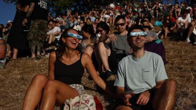 Two people smiling sat on a hill wearing eclipse glasses