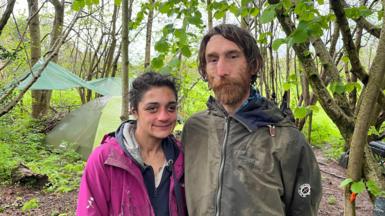 A man and woman stand in a wood in front of a green tent and tarpaulin tied between trees. The man has long brown hair, a brown beard and a green coat. The woman has black hair tied back, a navy hoodie and a pink coat.
