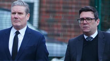 Two men dressed in dark suits - Keir Starmer on the left and Andy Burnham on the right - look grim-faced as they walk across a car park. Andy Burnham is wearing a black jumper underneath his suit jacket.