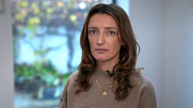 Claire Corkery, a woman with long hair, wearing a beige jumper and a gold necklace, standing in her dining room.