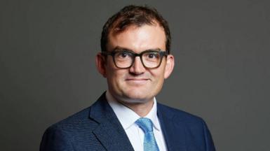Patrick Spencer wears a navy suit and stands in front of a plain background. He is smiling directly at the camera and is wearing large black-rimmed glasses. 