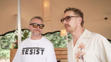 Simon Pegg and Benedict Cumberbatch stand side by side under a tent canopy. Pegg is smiling and laughing as he looks towards Cumberbatch. They are both wearing sunglasses. Pegg has on a white T-shirt which says 'Resist' in upper case, black letters. Cumberbatch has on a cream, short-sleeved shirt with a floral design on one side.