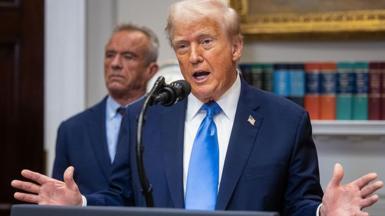 President Donald Trump stands in front of a microphone in the Oval Office at the White House, wearing a bright blue tie, white shirt and dark blue suit. He is speaking and holding both hands up. In the background are blurred books and a gold picture frame, with his Health Secretary Robert F Kennedy Jnr standing behind him.