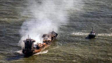 Smoke billows from a rust-coloured tanker ship floating in the sea. A tugboat floats nearby.
