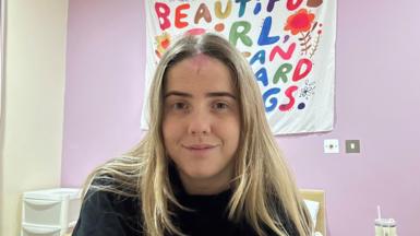 Rachel, who has blonde hair and is wearing a black jumper, sits on a bed with a set of portable draws beside it. A white banner on a purple wall behind her is partly obscured by her head, but the colourful letters on it appear to include the words 'Beautiful girl'