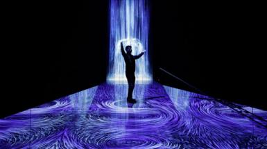 A person, in silhouette, poses at a sound and light installations. Blue light to resemble rain fall and puddles is projected in a black room