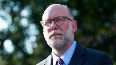 Russell Vought speaks to media outside wearing his glasses, a striped tie, button-down shirt and dark suit jacket