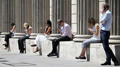 Five people sit along a low wall in the sunshine looking at their mobile phones, while a man stands at the right-hand side speaking to someone on the phone