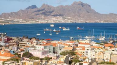 View over Mindelo, Sao Vicente, Cape Verde showing buildings with terracotta-coloured roofs in front of the blue ocean, which has several boats in it. Behind the ocean is a grey mountain and the sky is light and blue.