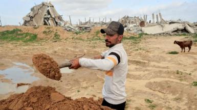 In Gaza, a Palestinian man shovels earth with a spade, with destroyed buildings and a goat in the background (10/01/26)