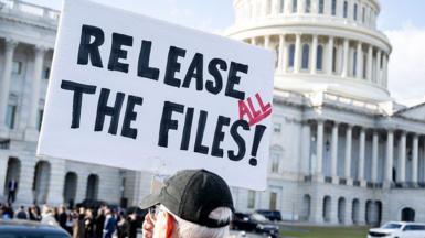 Man holding a sign that says "release all the files" with a black hat on standing outside the US Capitol on 12 November.