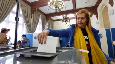 A woman with blond hair, wearing a yellow scarf and a blue jacket, is seen casting her vote at a polling station