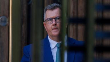 Jeffrey Donaldson, a man in a blue suit with short grey hair and glasses, seen through the bars of an entrance gate.