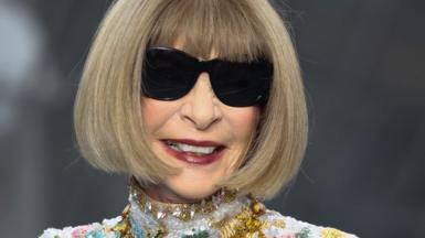 A close up headshot of Anna Wintour smiling, wearing sunglasses.  She has a bob haircut and is wearing a high collar embroidered dress studded with crystals.