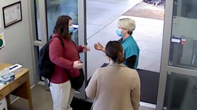 Three women stand talking in front of a sliding door at a hospital while wearing blue medical masks.