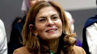 Close-up of Hind Kabawat, smiling. She has shoulder length brown hair and is wearing a smart, light brown coat.