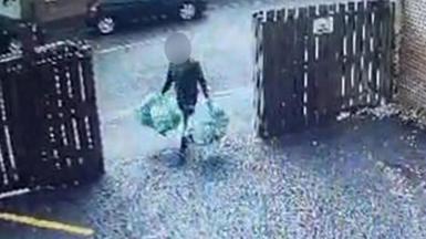 A man carrying four bags of rubbish, two in each hand. He is walking through an open gate into a driveway, with wooden fencing either side. His face is obscured. 